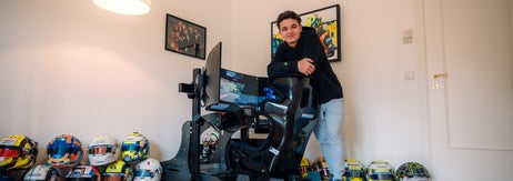 Person leaning on a racing simulator in a room with multiple racing helmets on the floor and framed racing memorabilia on the walls
